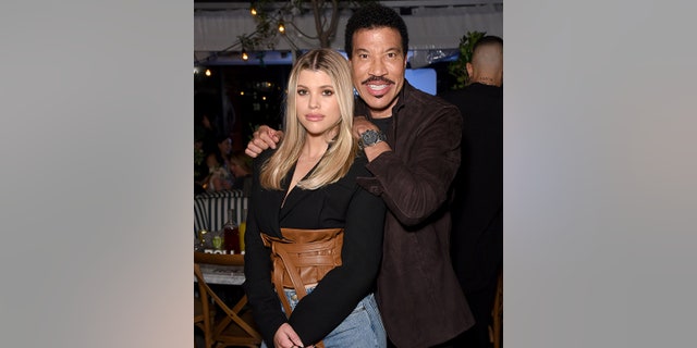 Sofia Richie in a black shirt and brown leather waist top soft smiles as her father Lionel Richie leans against her