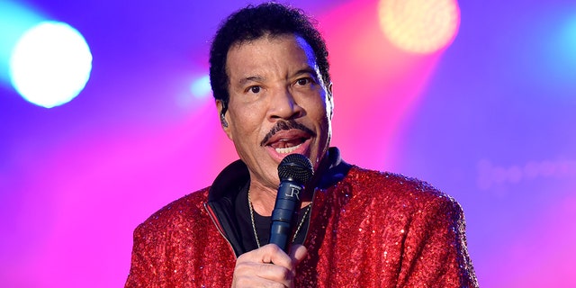 Lionel Richie speaks into the microphone on stage wearing a sparkly red jacket