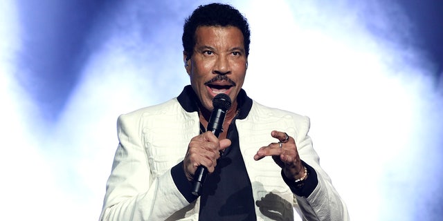 Lionel Richie in a white jacket and black shirt underneath performs on stage in front of a blue/white background