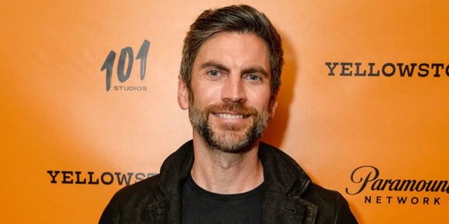 Wes Bentley in black smiles in front of an orange backdrop for a "Yellowstone" premiere