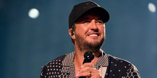 Luke Bryan in a patterned dark shirt and black cap looks up to his left while holding a microphone