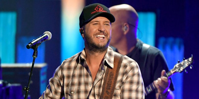 Luke Bryan in a checkered shirt and black cap plays the guitar at the ACM Awards