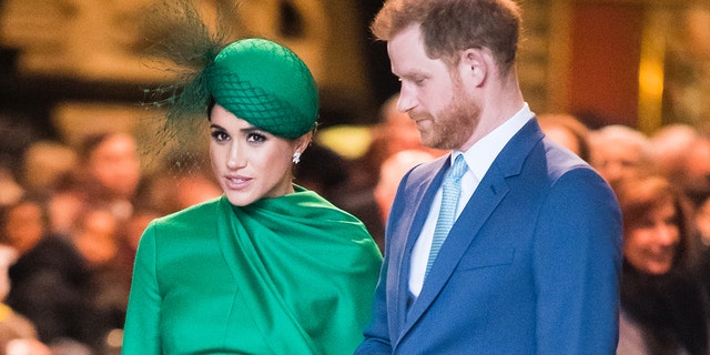 A close-up of Meghan Markle wearing a green dress and a matching hat standing next to Prince Harry in a blue suit and tie