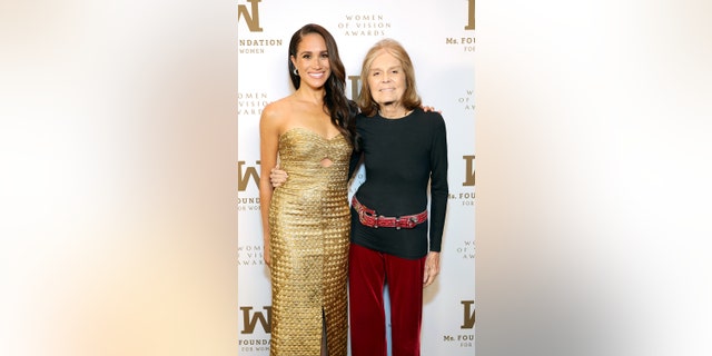 Meghan Markle in a gold strapless dress posing with Gloria Steinem who is wearing a black long sleeved shirt and burgundy pants