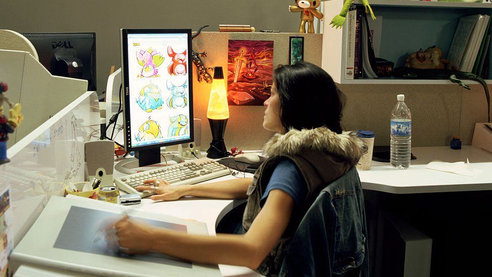 An artist using a drawing tablet on a computer, sat in an office. Her desk has a lava lamp as well as a screen on which six Neopets are visible.
