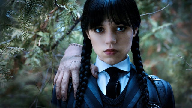 Wednesday Addams, clad in schoolgirl uniform and standing in a spooky pine forest, stares directly at the camera, with big dark eyes and a tough-girl pout. The disembodied hand named Thing perches on her shoulder, affectionately.