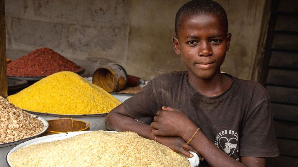 A young boy poses next to some rice in his shop at Ajara Market