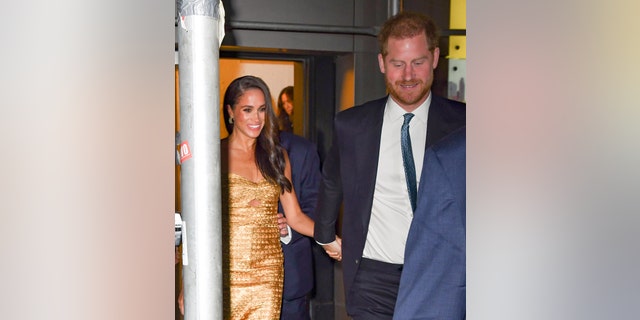 Meghan Markle wearing a gold dress being accompanied by Prince Harry in a navy suiit and white shirt