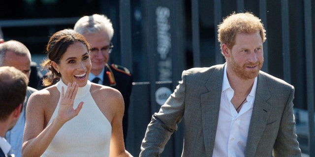 Meghan Markle waves in a white halter top and light pink pants while holding Prince Harry's hand, who sports a gray suit