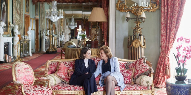 Lady Violet Manners in a navy blazer and pants with a white turtleneck sitting on a pink couch with her mother wearing a light blue blazer and a matching skirt.