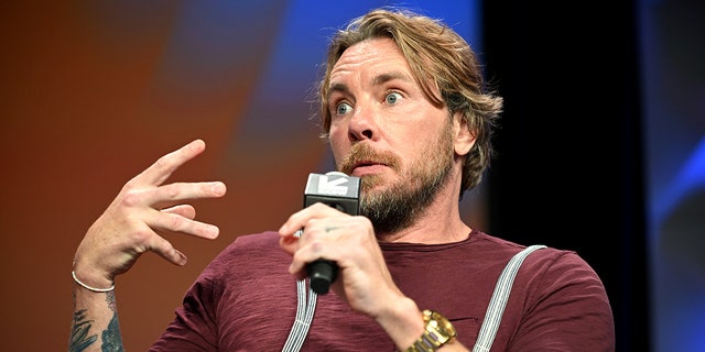 Dax Shepard in a maroon shirt and suspenders sits and gesticulates with his hand while talking into a microphone at SXSW
