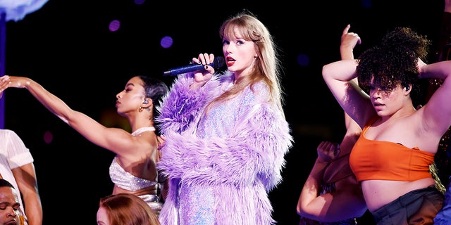 Taylor Swift sings on stage during "The Eras Tour" in a purple sparkle dress and feather lavender jacket