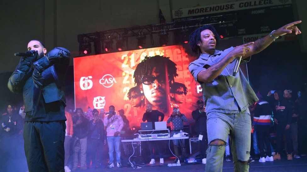 Drake and 21 Savage perform in Atlanta Georgia. They're in front of a large, red-hued picture of 21 Savage, showing his face from three different angles, with his name printed repeatedly behind him. Drake is wearing a black bomber-style jacket and thick gloves. His face is mostly obscured as he sings into the microphone. 21 Savage is wearing a short-sleeved shirt and shorts, and pointing at the crowd.