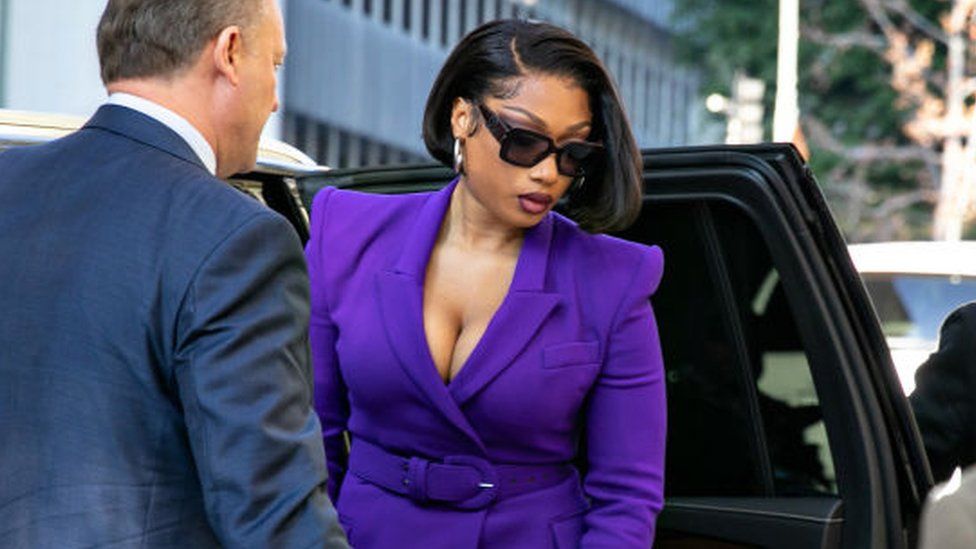 Megan Thee Stallion is helped out of a black Range Rover type vehicle by a minder as she arrives at court in Los Angeles. She's smartly dressed in a purple, belted jacket with a deep neckline. Her shoulder-length hair is straight and parted so one side covers one of her large silver hoop earrings. She's looking down at the ground through a pair of large, black sunglasses.