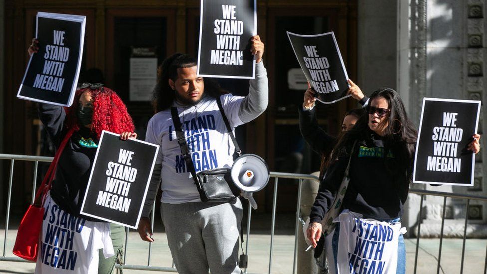 Four Megan Thee Stallion supporters stand outside a Los Angeles courthouse. They're each holding a sign with "We Stand With Megan" written in white block capitals on a black background. One, with a megaphone slung over his shoulder, wears a white sweatshirt with the same slogan printed in blue. Three of the protesters stand in front of a metal security railing, while the fourth - who stands behind a fellow protester, is on the other side of it.