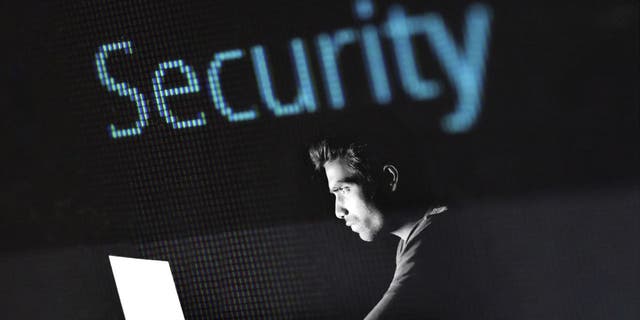 A man sitting in the dark, looking at a laptop, black and white filer, with the word "security" above him in blue