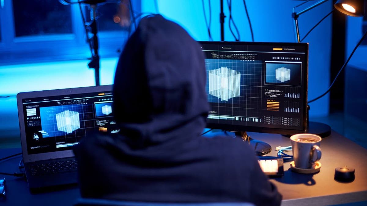 Hooded person looking at his computer monitors.
