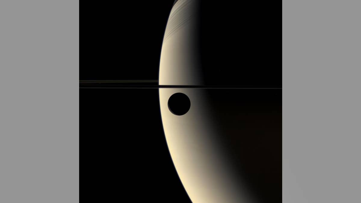 Saturn in the background with Rhea, one of the planets moons, seen in the foreground. A small thing line of Saturn's rings can also be seen