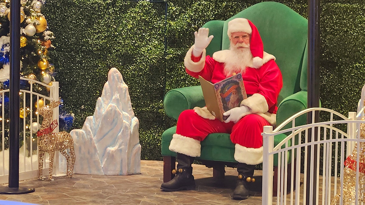 Santa Claus sitting in a big green chair in front of bushes, next to Christmas decor, with a book, waving