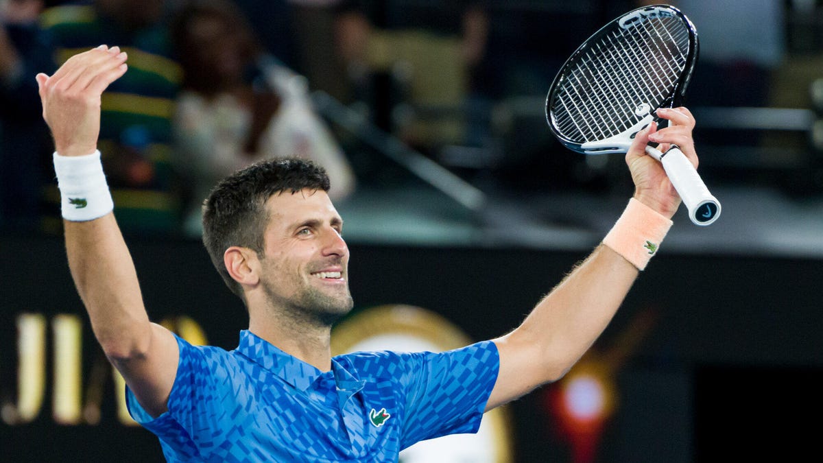 Tennis star Novak Djokovic celebrating with both arms raised, holding a raquet in his left hand.