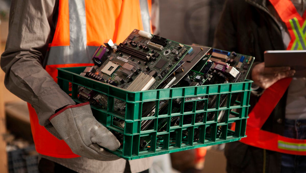 A worker in protective gear carries a container full of old motherboards and other computing parts.