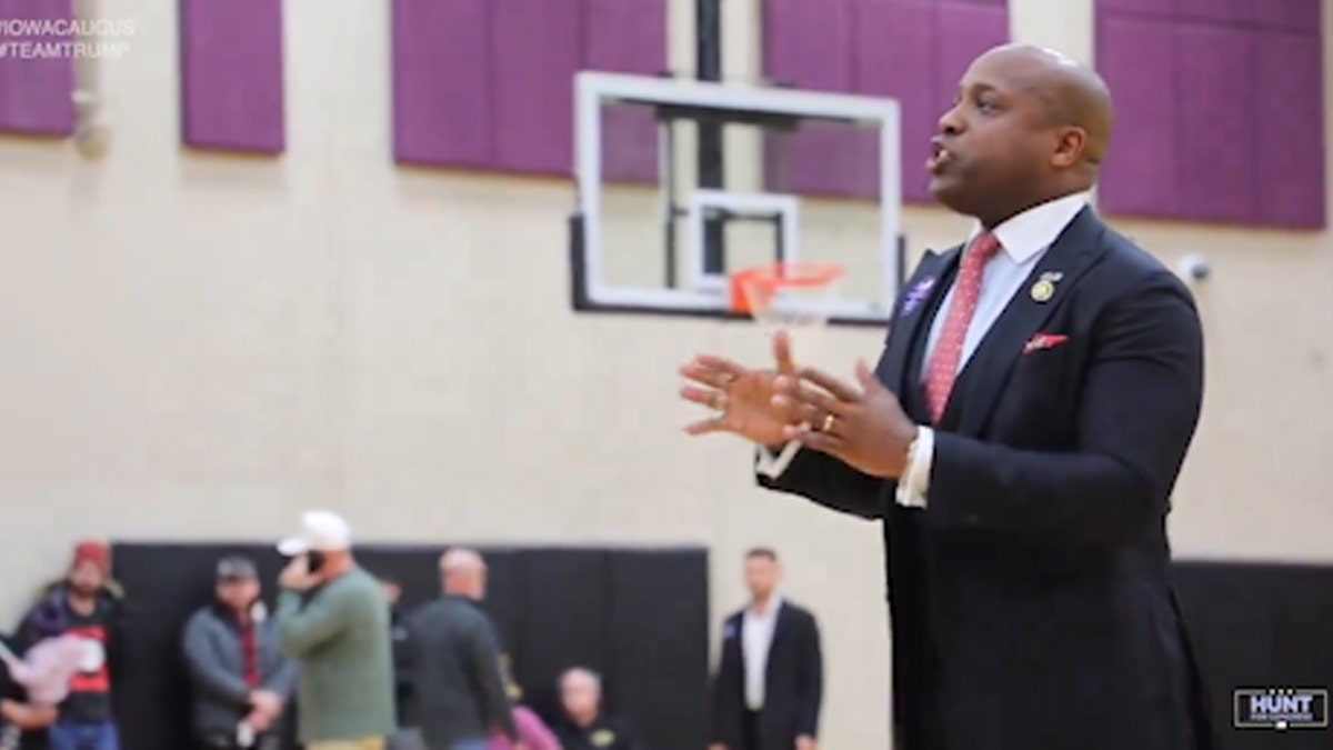 Rep. Wesley Hunt speaking, black suit, red polka dot tie, men in background, in school gym with reddish/purple tiles on the wall and basket ball net and hoop