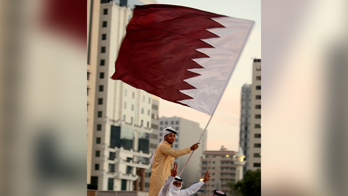 Qatari man waving Qatar flag, dark red and white, other Qatari man sitting and giving peace signs