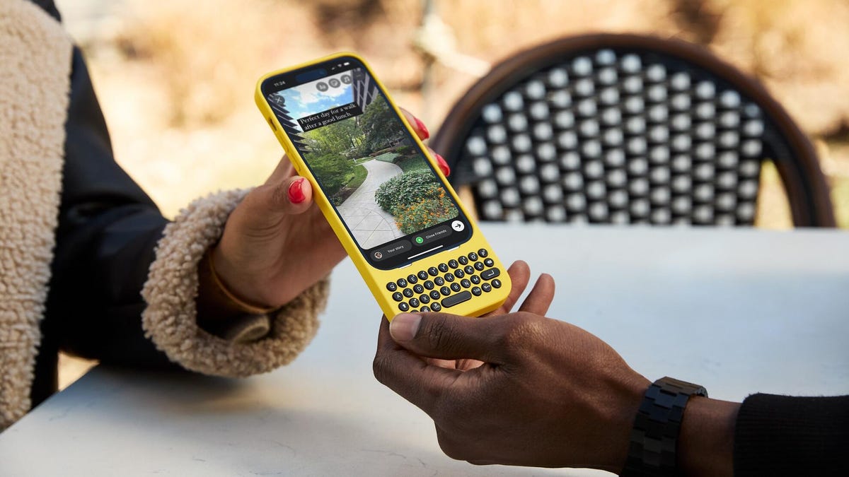 A person hands their iPhone to another to hold -- the phone is wrapped in the Clicks case-and-keyboard, which is bright yellow with stylish black keys.