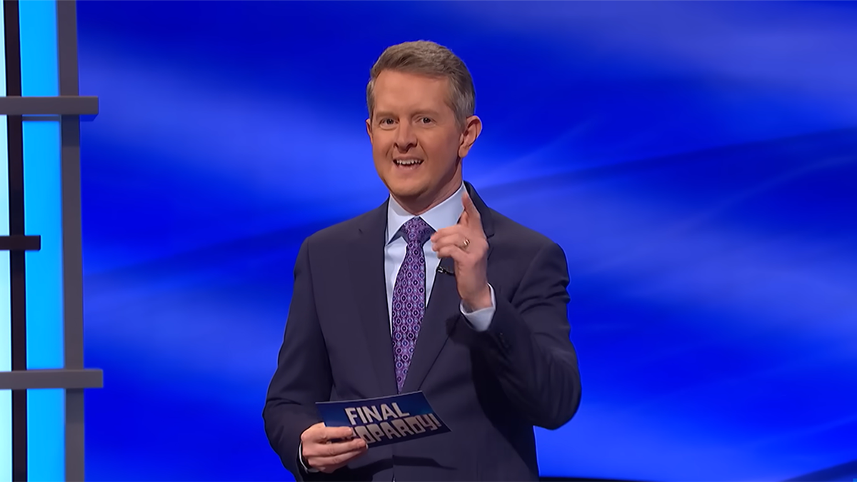 Ken Jennings in a navy suit with a light blue shirt and purple suit on the set of 'Jeopardy!' pointing at the camera