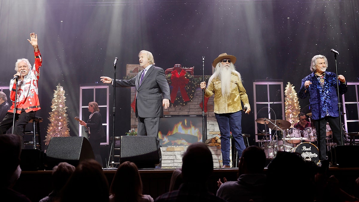 Joe Bonsall in a red shirt raises his hand to the crowd in Nashville, Tennessee from his stool while performing alongside The Oak Ridge Boys