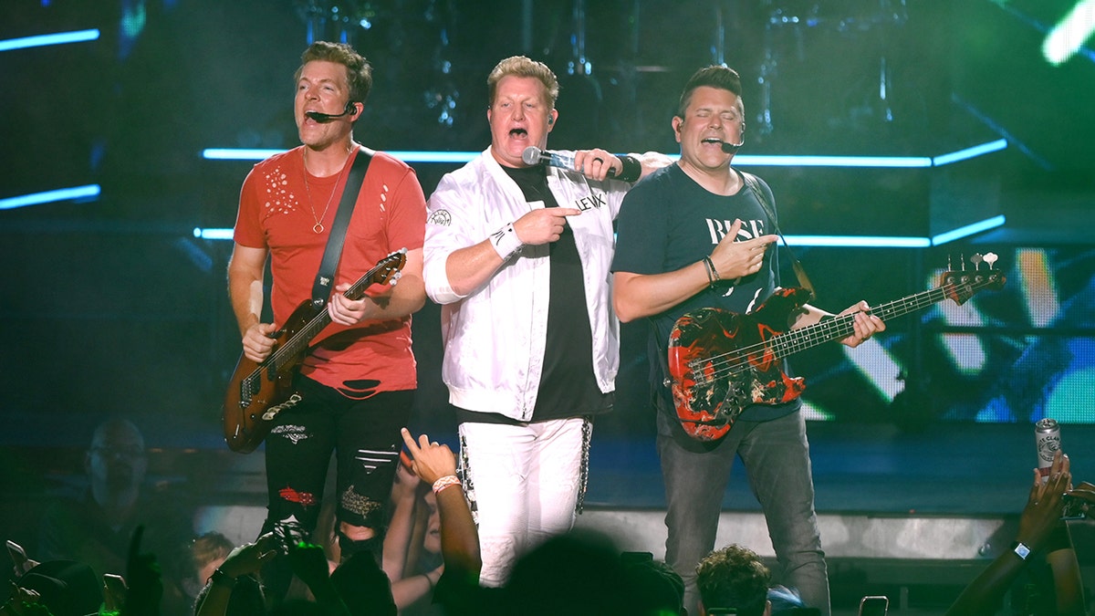 Joe Don Rooney in a red shirt, Gary LeVox in a white jacket and pants and Jay DeMarcus in a dark shirt perform together on stage in Tennessee