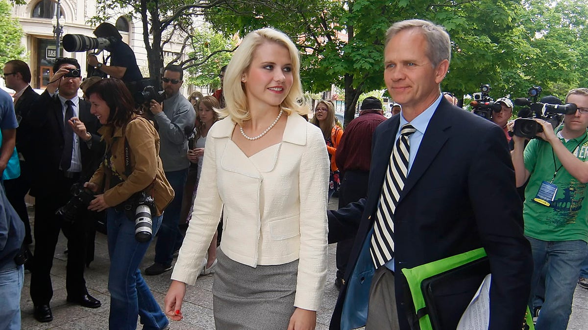 Elizabeth Smart in a white blazer and pearls walking next to her father in a suit and tie