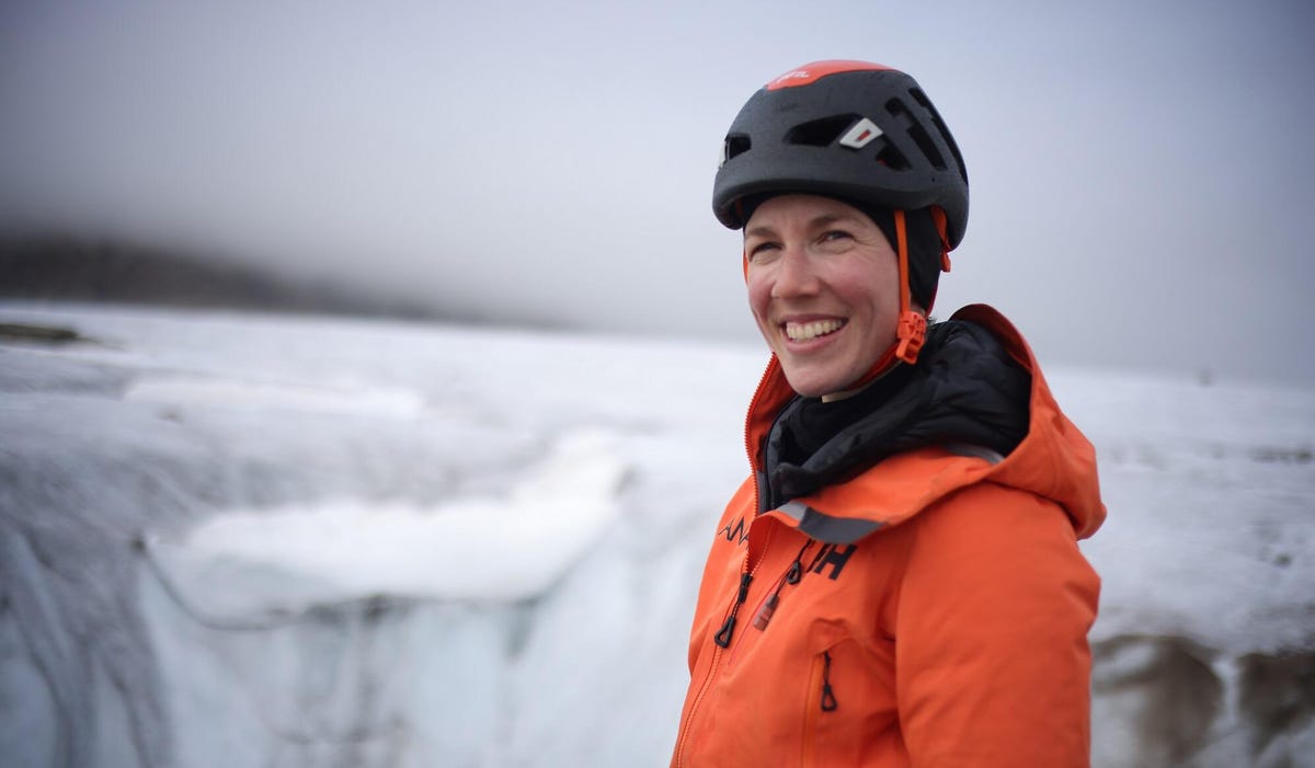 A woman smiles while wearing a black safety helmet and orange winter coat in front of a background of ice and glaciers.