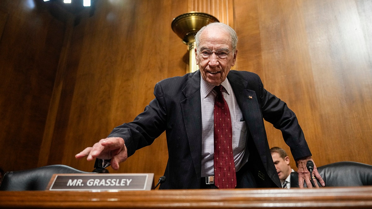 Chuck Grassley in a dark suit, red tie, reaching out right hand on table, left hand on chair
