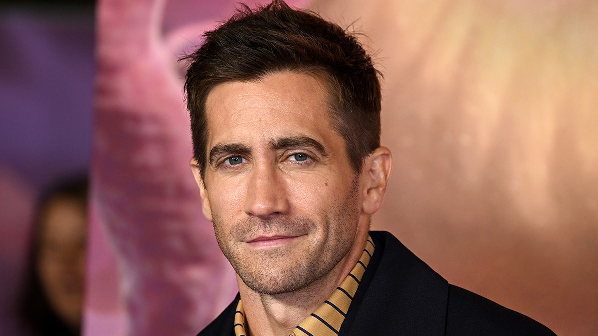 Jake Gyllenhaal in an orange stiped shirt and black jacket on the red carpet soft smiles