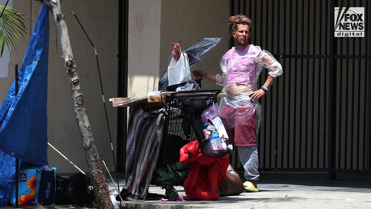 A homeless man in a clear plastic poncho stands next to a shopping cart