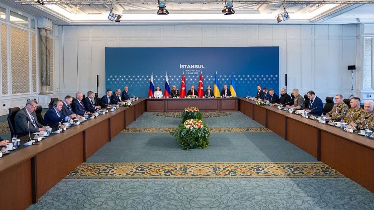 Delegations from Russia and Ukraine sit across from each other at a long U-shaped table during peace talks in Istanbul, Turkey, with Turkish officials at the head table and national flags from Russia, Turkey, and Ukraine displayed behind them. The room is elaborately decorated with carpeted floors, ornate wallpaper, and overhead lighting.