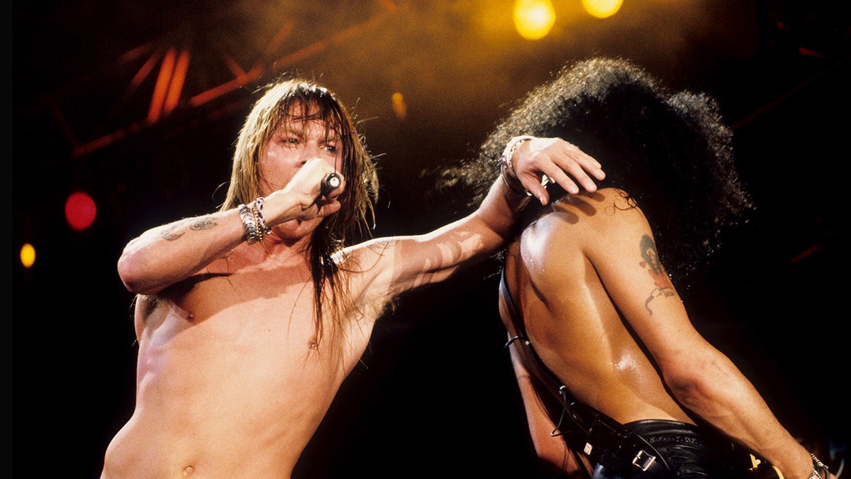 Axl Rose leaning on Slash while singing on stage.