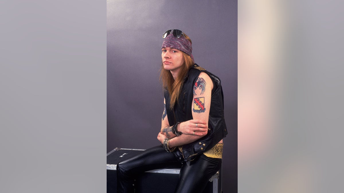 Axl Rose wearing leather pants and a black vest with a purple bandana showing off his tattoos.