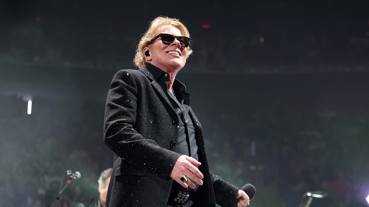 Axl Rose wearing all black with sunglasses on a stage smiling.