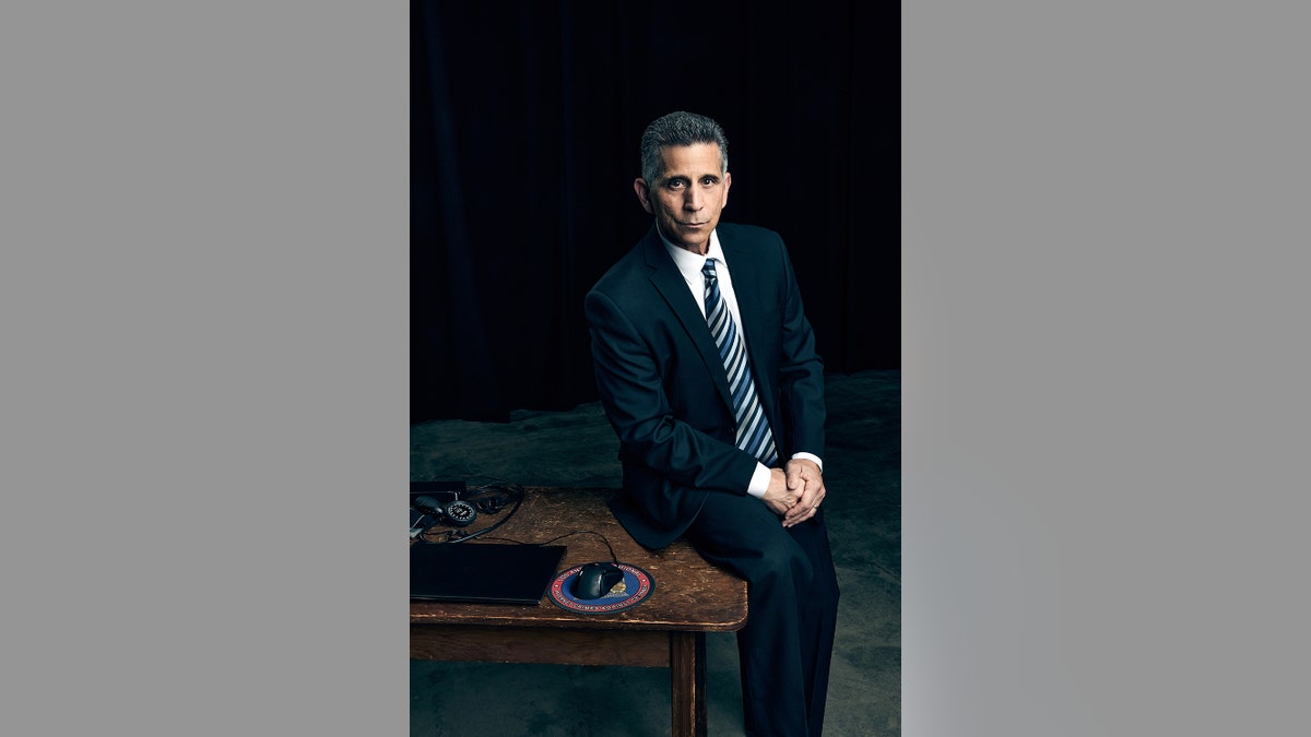 George Olivo in a dark suit sitting on a desk.