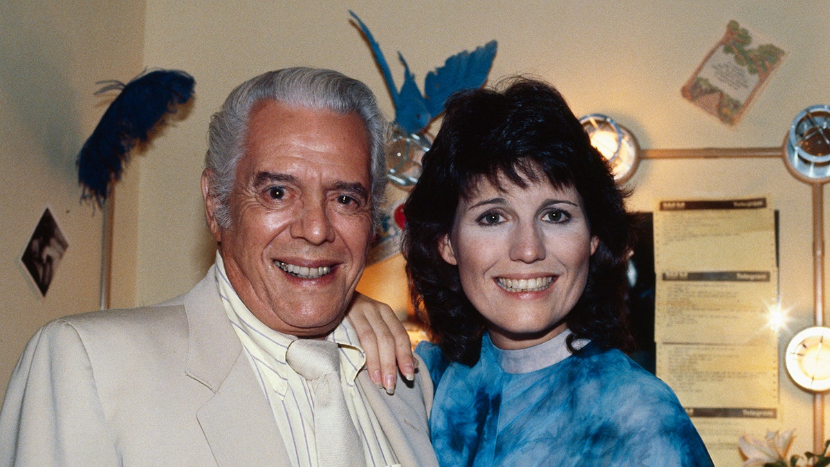Lucie Arnaz in a bright blue shirt smiling and leaning on her father who is wearing all beige.