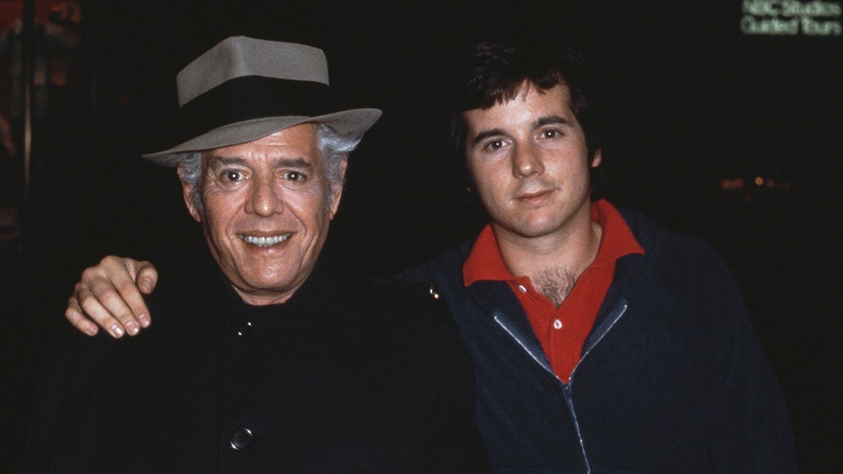 Desi Arnaz Jr. with his arm around his smiling father as they wear matching black outfits.