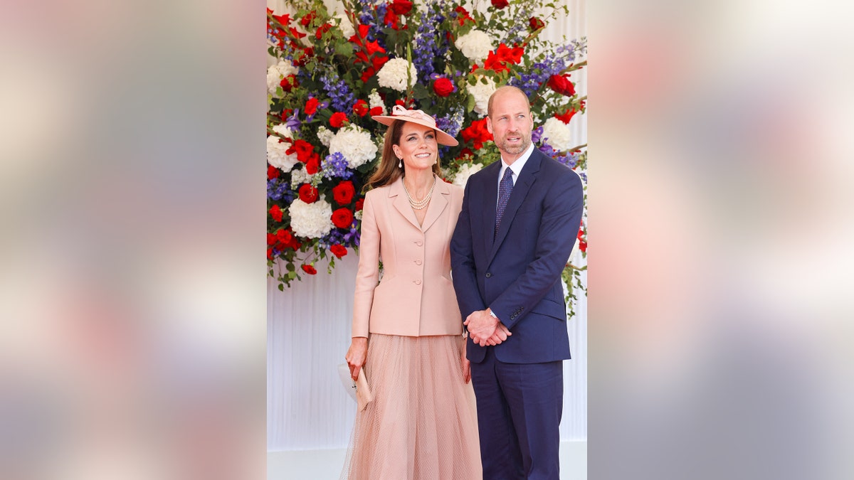 Kate Middleton wearing a pink Dior suit standing next to Prince William in a blue suit with flowers behind them.