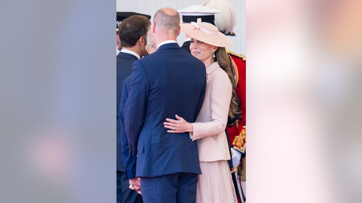 Kate Middleton holding on to Prince William who is wearing a blue suit as she wears a pale pink coat dress.