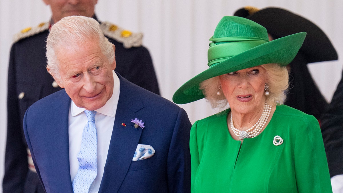 King Charles in a blue suit standing next to Queen Camilla in an all green coat dress.