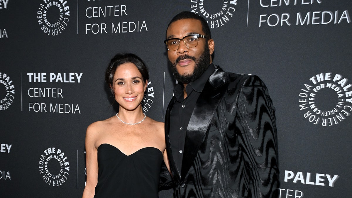 Meghan Markle wearing a strapless sweetheart cut black dress standing next to Tyler Perry wearing all black.