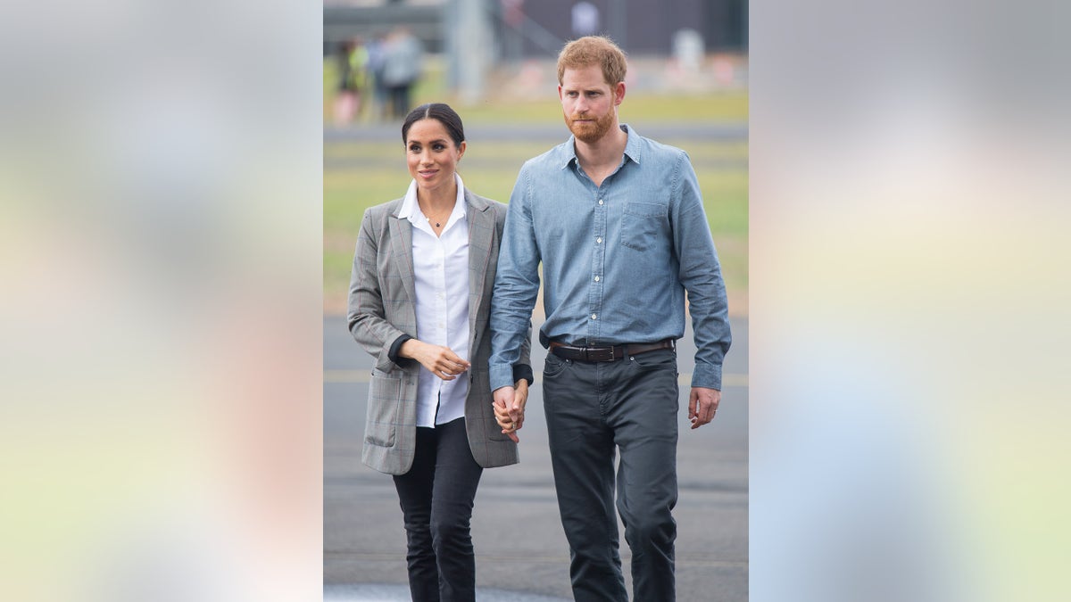 Meghan Markle wearing a white blouse and a grey jacket walking alongside Prince Harry wearing a blue shirt and dark blue pants.