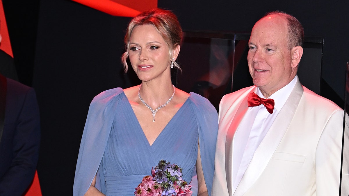 Princess Charlene wearing a flowy blue dress and holding a bouquet of flowers standing next to Prince Albert wearing a white suit with a red bowtie.