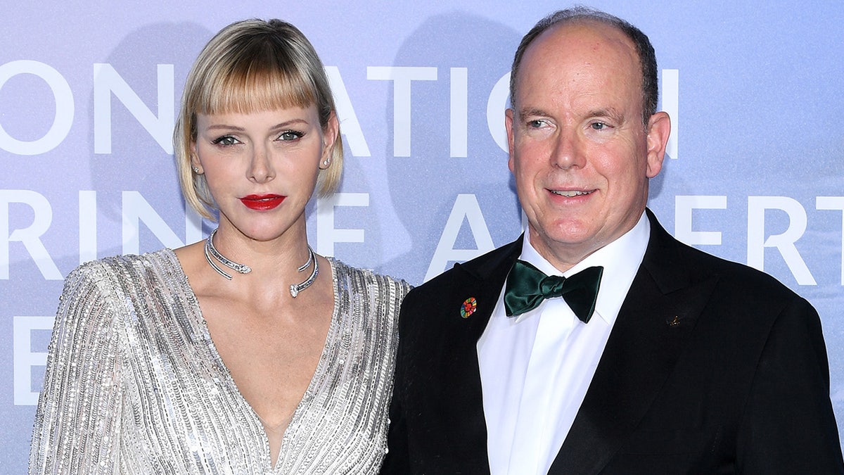 Princess Charlene wearing a sparkling silver dress standing next to Prince Albert in a tux and bowtie.